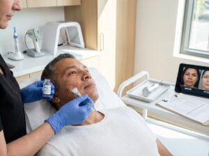 Dermatologist applying salicylic acid peel to an adult patient with acne in a modern clinic, with microneedling, laser and LED devices visible in the background