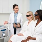 Clinician in a modern dermatology clinic comparing a BBL Hero device and an IPL handpiece with two diverse patients, tablet showing skin before and after images